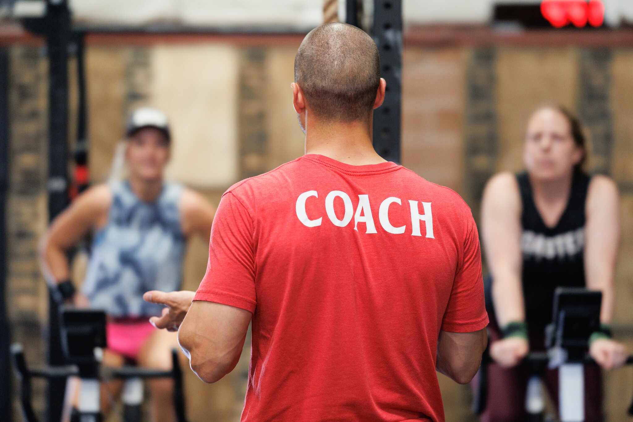 Treinador visto de costas, com a palavra COACH estampada na camisa, orientando alunas em uma academia