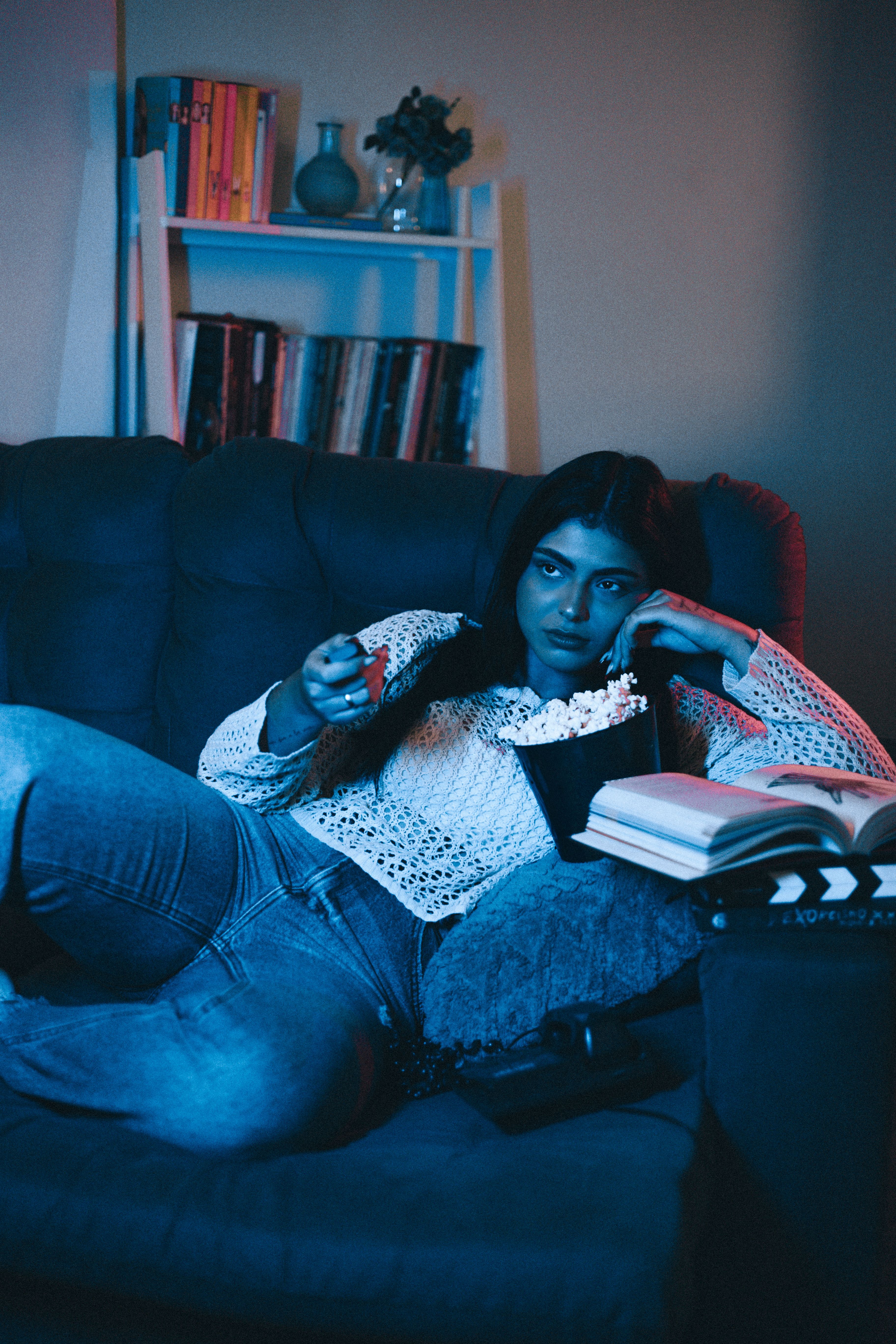 Mulher deitada no sofá assistindo TV e comendo pipoca, com livros ao lado