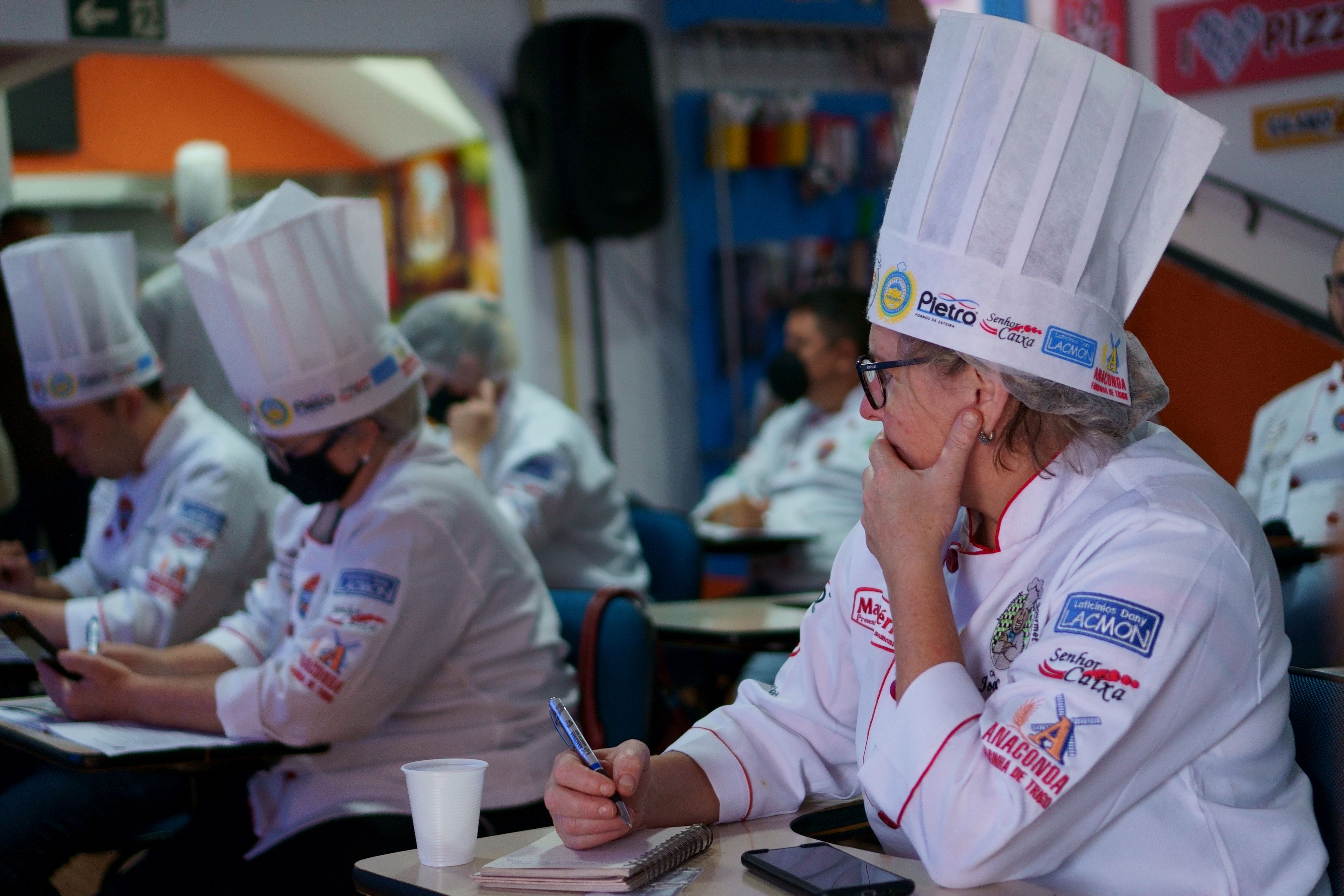 Cozinheiros de chapéu alto, um deles pensativo, anotando algo em um bloco de notas