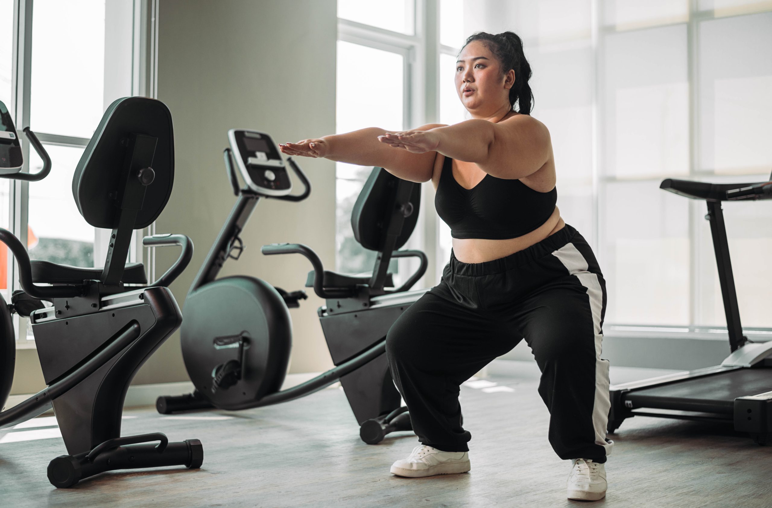 Mulher treinando agachamento numa academia, cercada de equipamentos cardiovasculares